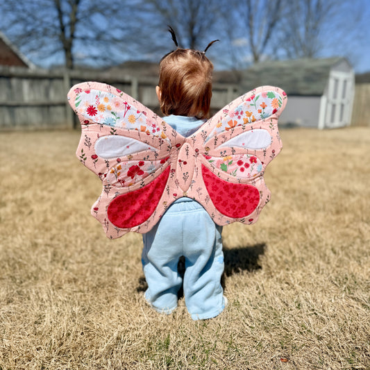 Child wearing a butterfly costume with floral wings outdoors on a grassy area.