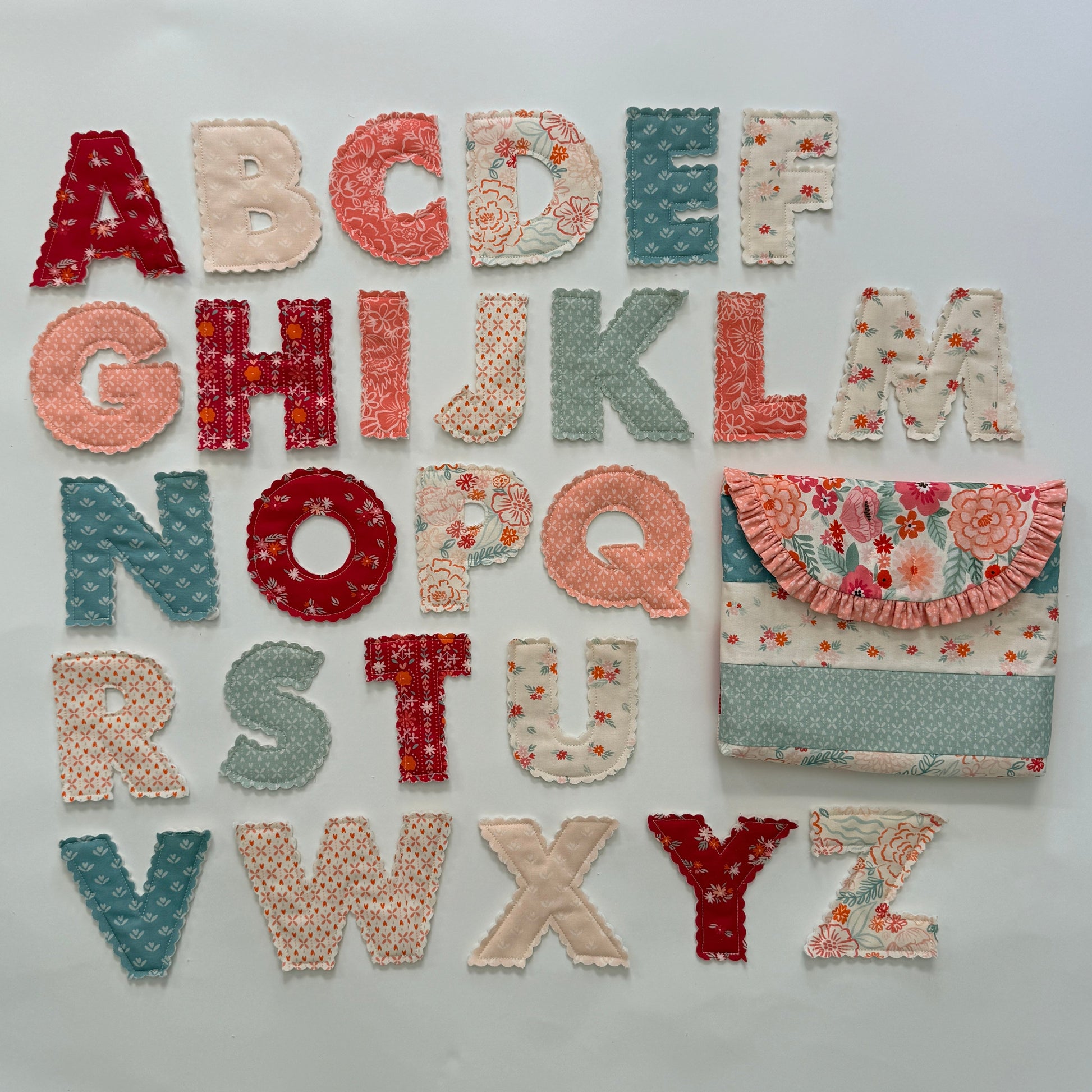 A set of handmade fabric alphabet letters arranged on a white background, alongside a matching fabric pouch. The letters and pouch feature various patterns including floral prints, polka dots, and textured knit fabrics in muted red, peach, sage green, and cream colors.
