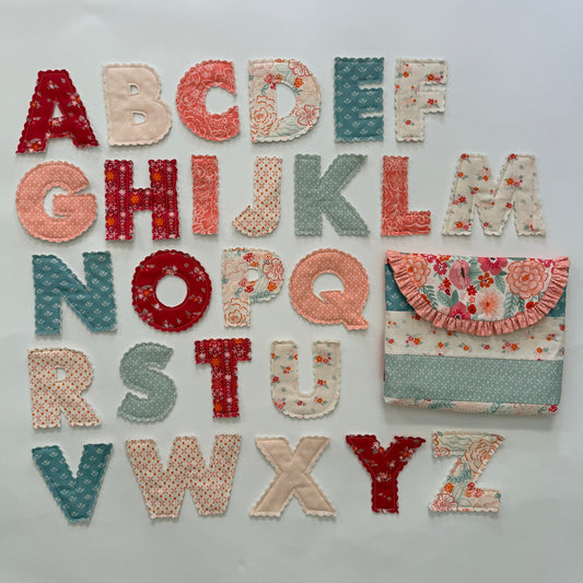 A set of handmade fabric alphabet letters arranged on a white background, alongside a matching fabric pouch. The letters and pouch feature various patterns including floral prints, polka dots, and textured knit fabrics in muted red, peach, sage green, and cream colors.