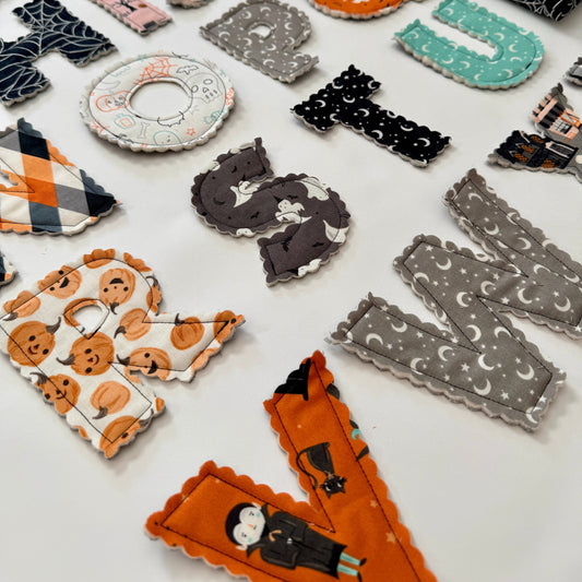 A collection of handmade, fabric-covered alphabet letters arranged on a white surface, featuring various Halloween-themed patterns like pumpkins, spiderwebs, ghosts, and witches.