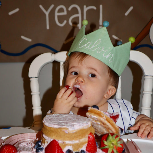 Child celebrating a birthday with a cake and birthday crown, sitting in a high chair.