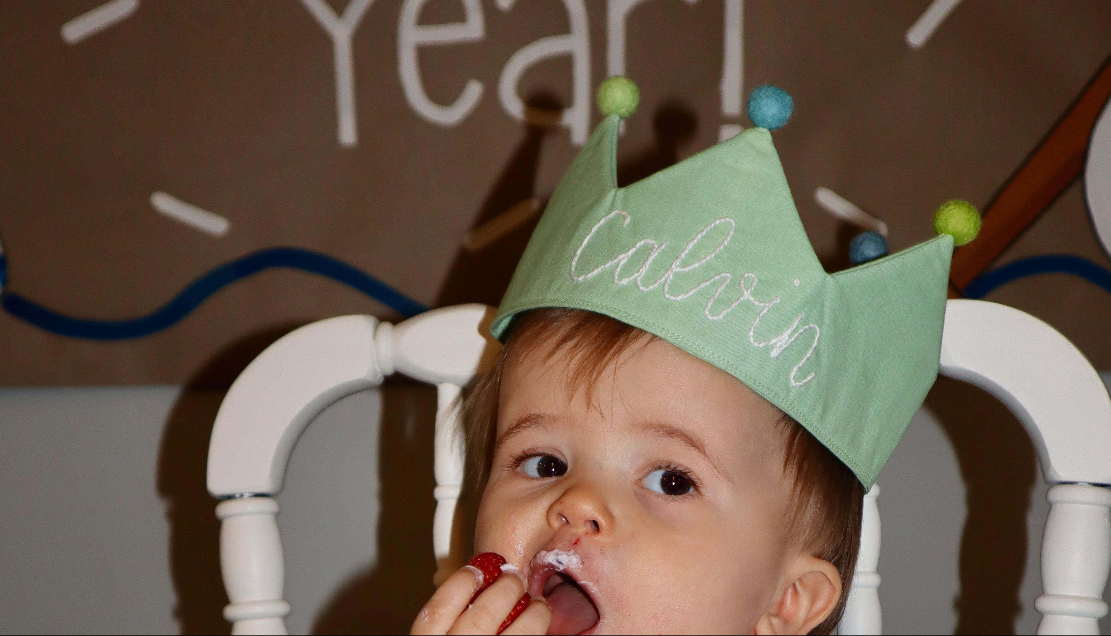 Child celebrating a birthday with a cake and birthday crown, sitting in a high chair.