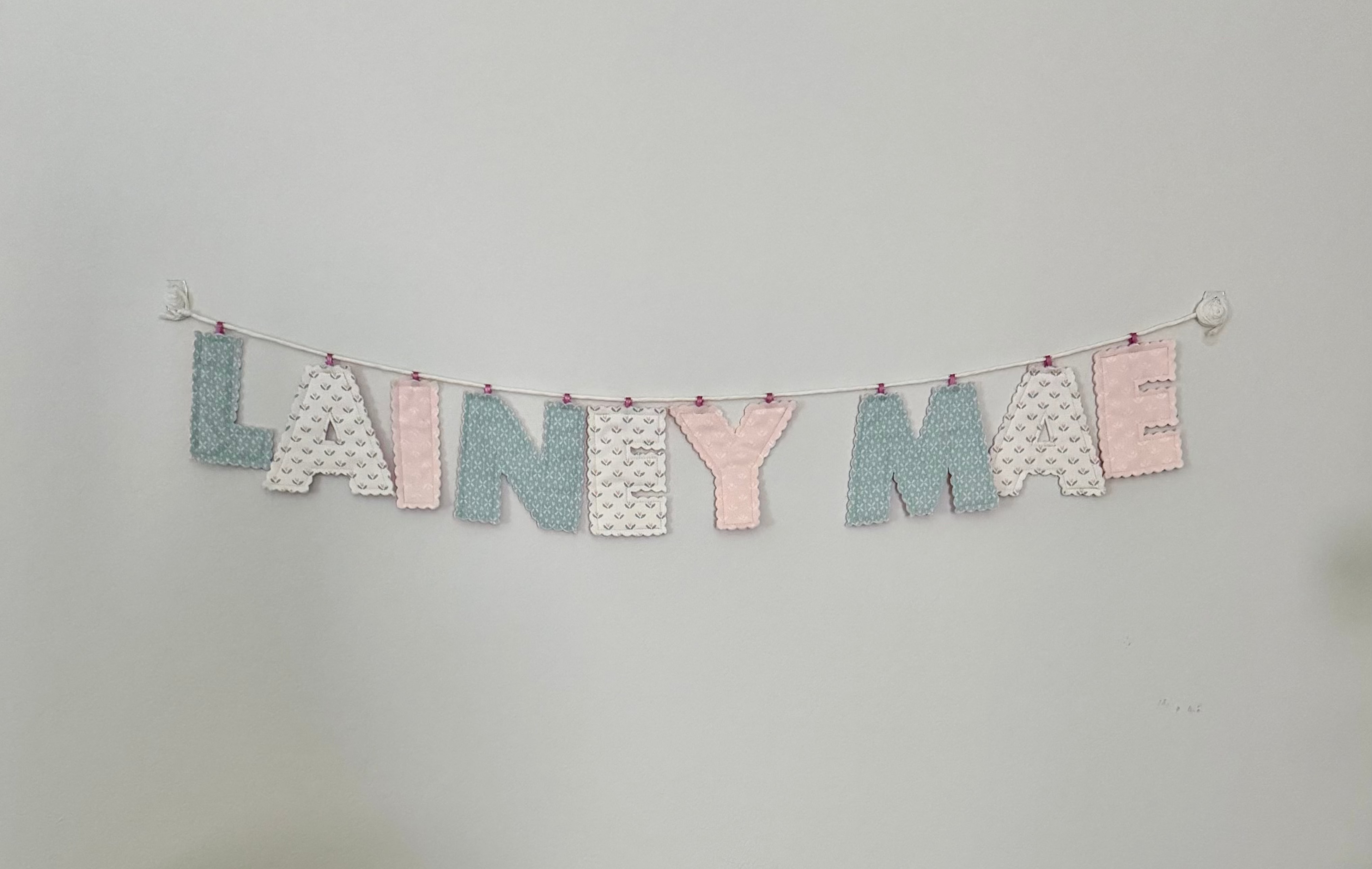 Wooden crib with floral bedding and a teddy bear, with a name banner above on a plain wall.